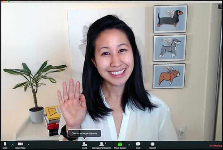 Smiling woman in video call, waving, with plant and dog art on wall background, perfect for virtual meeting or remote work.