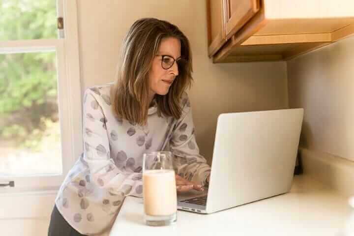 Woman working from home on laptop in kitchen, wearing glasses, with a glass of milk on counter. Remote work lifestyle.
