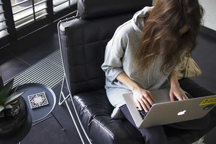 Woman in grey hoodie using laptop on a black chair beside a small table with a plant and notebook.