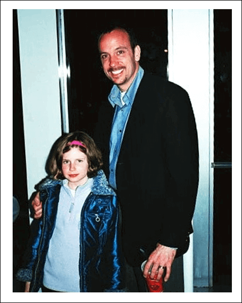 Father and daughter smiling indoors, man in suit holding drink, girl in blue jacket with pink headband.