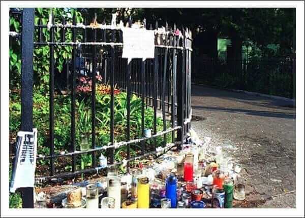 Memorial candles lined up along a black metal fence in a park setting, honoring a special remembrance occasion.
