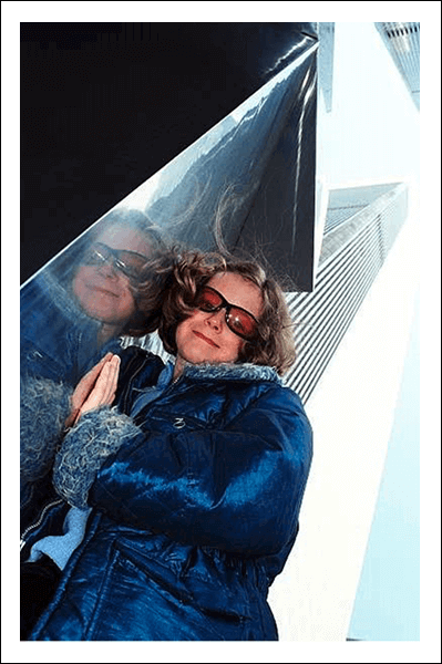 Person in blue jacket and sunglasses leaning on modern architecture with reflective surfaces in urban setting.