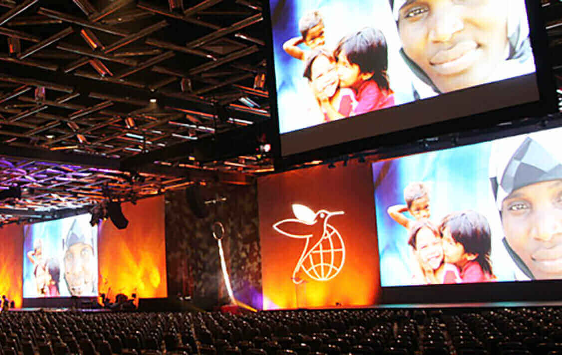 AV broadcast event with large screens displaying diverse faces and orange-lit stage, showcasing global unity theme.