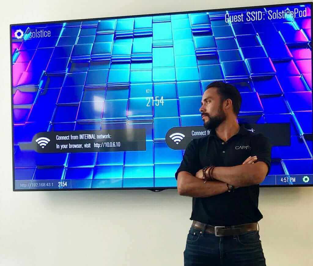 Man standing in front of a large digital screen displaying network connection info, wearing a black shirt, arms crossed.