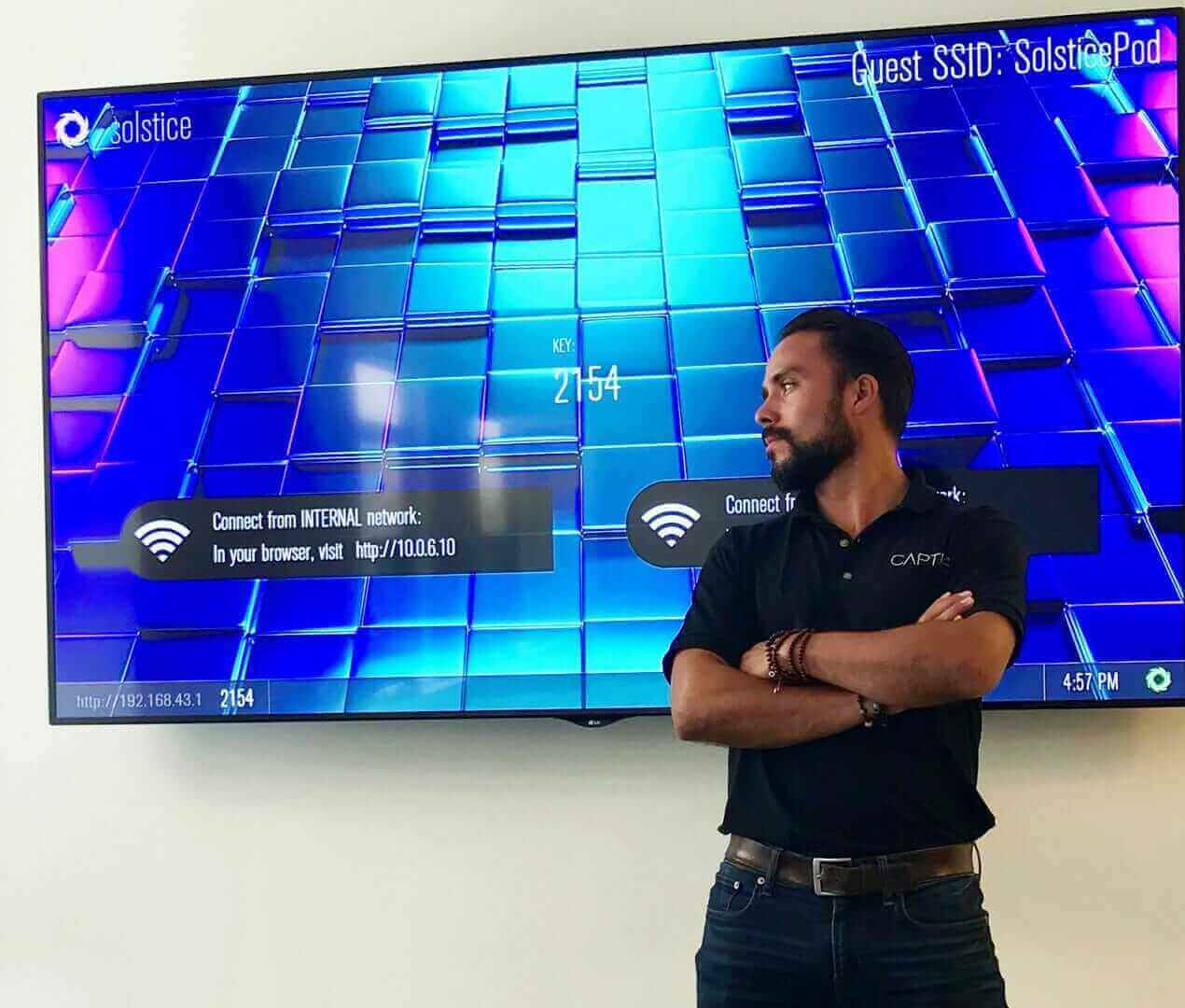 Man standing in front of a large digital screen displaying network connection info, wearing a black shirt, arms crossed.