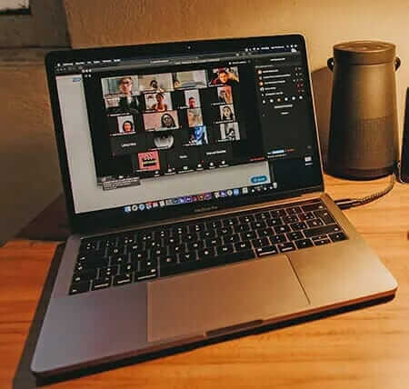 Laptop displaying virtual conference call, featuring multiple participants, on a wooden desk with a speaker nearby.
