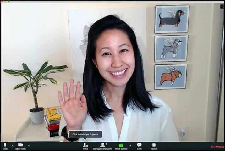 Smiling woman waving during video call, home office setting, plant and dog art in background, Zoom meeting interface visible.