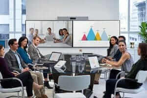 Modern office meeting with diverse team using video conference and laptops, discussing data charts on large screen.