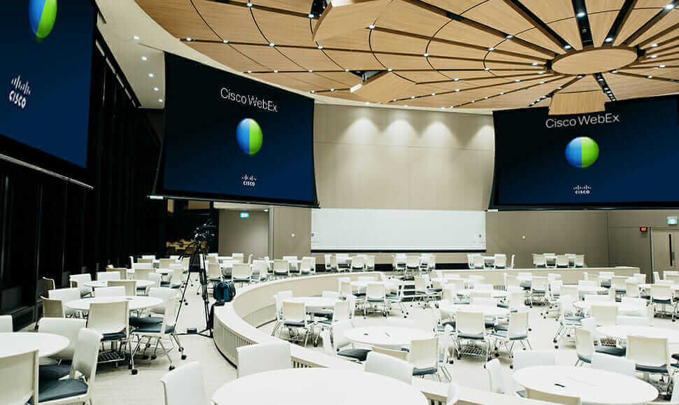 Modern conference room with circular seating, AV broadcast screens displaying Cisco WebEx, and a wooden panel ceiling.
