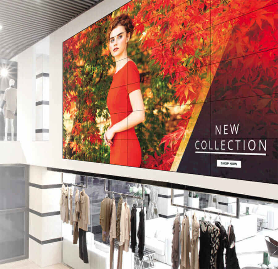 Fashion store interior with large digital screen displaying new collection ad featuring woman in red dress; clothing racks below.