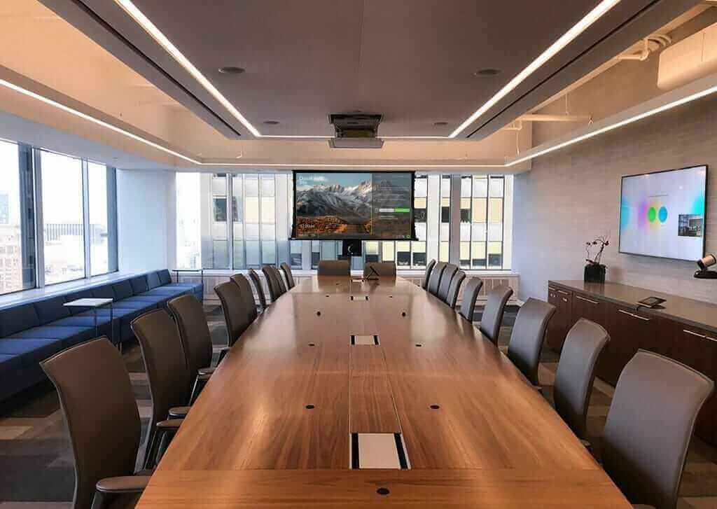 Modern conference room with long wooden table, leather chairs, digital screens, and large windows offering city view.