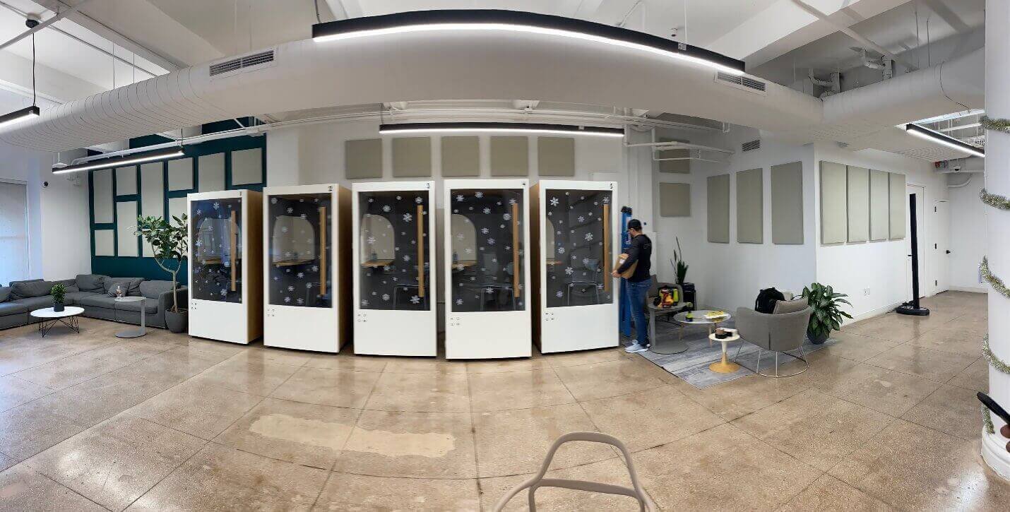 Modern office space with soundproof phone booths, lounge area, plants, and a person adjusting equipment, showcasing work environment.